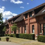 Backsteingebäude mit markanten Giebeln und gepflegtem Vorgarten bei blauem Himmel und Sonnenschein.