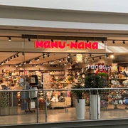 Nanu Nana Das Bild zeigt den Eingang eines Nanu-Nana-Geschäfts in einem Einkaufszentrum. Durch die gläserne Fassade sind viele bunte und dekorative Artikel zu sehen, die im Inneren des Ladens auf Regalen ausgestellt sind.