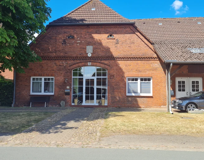 Dreppenstedt Ziegelgebäude mit großen Fenstern, gepflastertem Weg, seitlich ein Auto unter einem Carport.