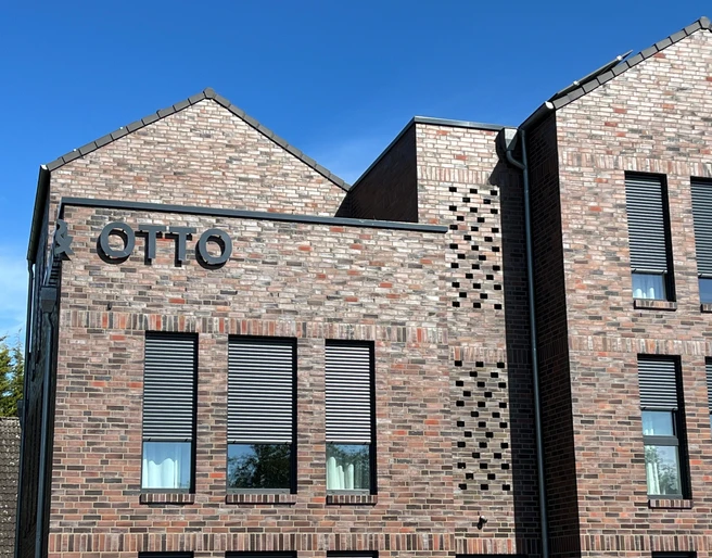 linda&otto Rotes Ziegelgebäude mit modernen grauen Fensterläden und großem Schriftzug "OTTO" an der Fassade.