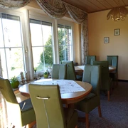 Pension Haus Marbeke Frühstücksraum.jpg Ein lichtdurchfluteter Frühstücksraum mit grünen Polsterstühlen und runden Holztischen.