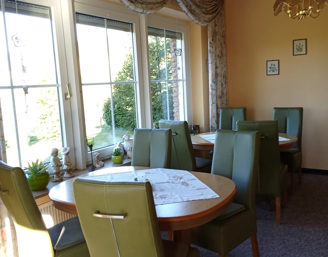 Pension Haus Marbeke Frühstücksraum.jpg Ein lichtdurchfluteter Frühstücksraum mit grünen Polsterstühlen und runden Holztischen.