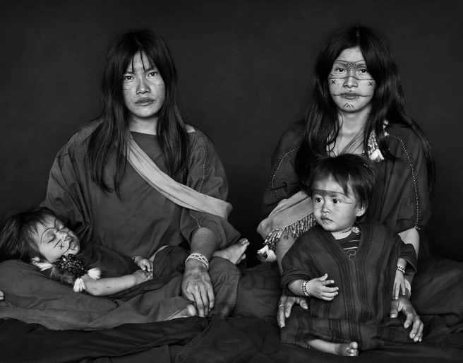 Amazonia Sebastiao Salgado Eine indigene Familie. Zwei Erwachsene sitzen zentral, während zwei Kinder auf ihnen ruhen. Die Gesichter sind mit traditionellen Mustern bemalt. Ein Ausdruck von Ruhe und Zusammenhalt dominiert die Szene.An indigenous family. Two adults sit in the center while two children rest on them. The faces are painted with traditional patterns. An expression of calm and cohesion dominates the scene.