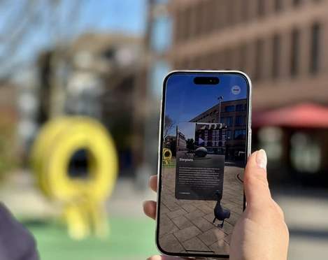 Stadtrallye.jpg Ein Smartphone wird draußen gehalten, das eine App mit Augmented Reality enthält, während im Hintergrund ein gelbes Kunstobjekt und Gebäudefronten sichtbar sind.