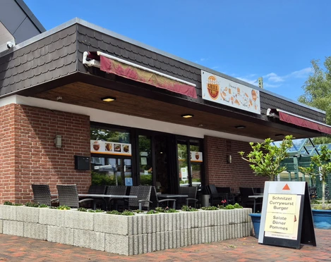 Aussenansicht Rasteder Grill Ein kleines, rotes Backsteingebäude mit Terrasse und überdachten Sitzplätzen wird von einem Schild mit Speiseangebot flankiert.