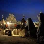 Bastei Erlebnis Steinreich Holztor mit Türmen und Fahnen, beleuchtet bei Nacht, umgeben von Bäumen und großen Steinen, vor einem tiefblauen Himmel.