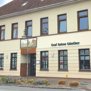 Restaurant Brassierie Graf-Anton-Günther Das Bild zeigt das zweistöckige Gebäude "Graf Anton Günther" in einer urbanen Umgebung. Die Fassade ist in Beige gehalten und das Dach mit roten Ziegeln gedeckt. Um das Gebäude sind Straßen mit Verkehrsschildern und einem Fußgängerüberweg zu sehen.