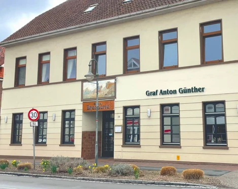 Restaurant Brassierie Graf-Anton-Günther Das Bild zeigt das zweistöckige Gebäude "Graf Anton Günther" in einer urbanen Umgebung. Die Fassade ist in Beige gehalten und das Dach mit roten Ziegeln gedeckt. Um das Gebäude sind Straßen mit Verkehrsschildern und einem Fußgängerüberweg zu sehen.