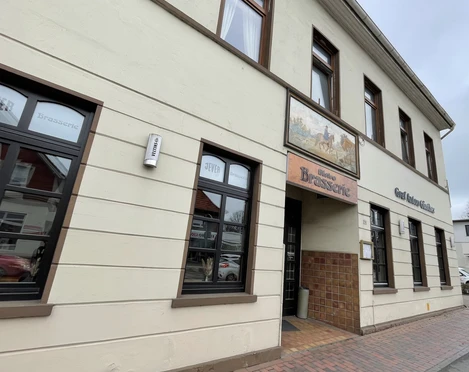 Restaunrant Brasserie Graf-Anton-Günther Das Bild zeigt die Außenfassade eines zweistöckigen Gebäudes, das als "Brasserie" ausgewiesen ist. Die Wände sind beige mit braunen Fensterrahmen. Über dem Eingang hängt ein farbiges Schild mit einem Pferdemotiv. Neben der Tür befindet sich ein Leuchtkasten mit der Aufschrift "Essen".