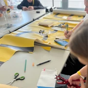 re:create Workshop <p>Menschen basteln an einem Tisch mit buntem Papier und Werkzeugen in einer hellen Umgebung.</p>