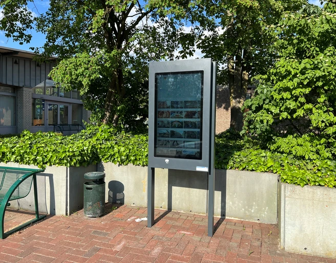 Infoterminal Eystrup Ein modernes Infoterminal steht auf einem gepflasterten Platz, umgeben von Bäumen und Büschen.