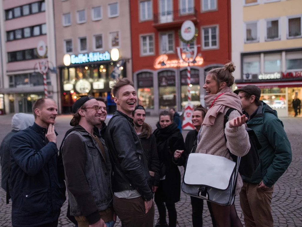 Eine fröhliche Gruppe erkundet lächelnd die Altstadt von Köln, umgeben von historischen Gebäuden und typisch kölschen Brauhäusern.A cheerful group smiles as they explore Cologne's old town, surrounded by historic buildings and typical Cologne breweries.