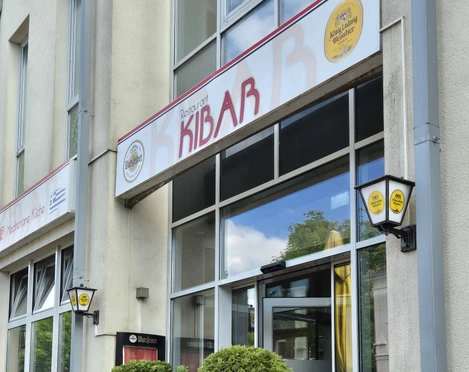 Restaurant Kibar Vor einem modernen Gebäude mit hohen Glasfenstern steht das Restaurant Kibar. Zwei Topfpflanzen flankieren den Eingang, daneben befinden sich draußen Tische und Stühle für Gäste.