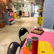 Museum des Schmerzes und der Liebe Im Vordergrund steht ein rosafarbener Tisch mit bunten Bechern, einer Chipstube und einem Schachbrett.In the foreground, there is a pink table with colorful cups, a tube of chips, and a chessboard.
