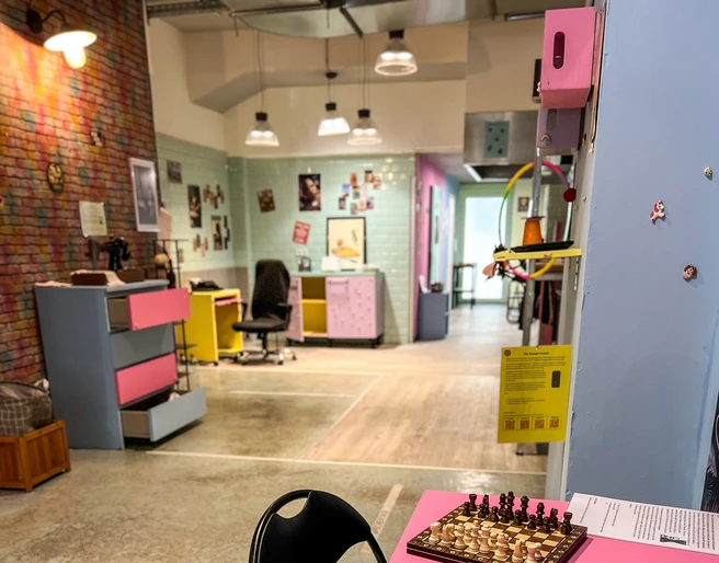 Museum of pain and love Im Vordergrund steht ein rosafarbener Tisch mit bunten Bechern, einer Chipstube und einem Schachbrett.In the foreground, there is a pink table with colorful cups, a tube of chips, and a chessboard.