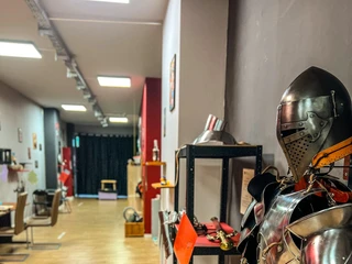 Museum of pain and love Das Bild zeigt eine detailreiche Ritterrüstung aus glänzendem Metall, die in einem modern gestalteten Museumsraum ausgestellt ist. Im Hintergrund sind verschiedene Exponate und Möbel auf einem hellen Holzboden zu sehen.The image shows a detailed knight's armor made of shiny metal, displayed in a modernly designed museum space. In the background, various exhibits and furniture are visible on a light wooden floor.