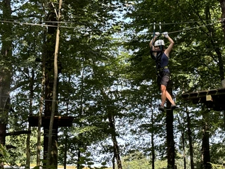 Kletterwald Koenigstein Kletterwald Königstein: Person in schwarzer Kleidung und Helm balanciert auf einem Seil im Kletterpark, umgeben von grünen Bäumen bei sonnigem Wetter.