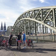 Colonia Aktiv guided bike tours in Cologne Gruppe von Menschen mit roten Fahrrädern auf einer Brücke in Köln, Dom im Hintergrund.Group of people with red bicycles on a bridge in Cologne, with the Cologne Cathedral visible in the background.