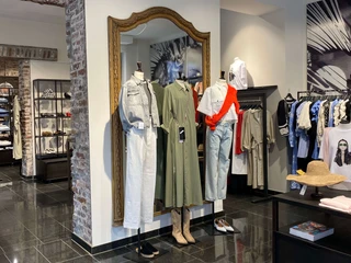 Simon und Renoldi  Das Bild zeigt eine Boutique mit modernem Interieur. Zwei Schaufensterpuppen präsentieren modische Kleidung: ein khakifarbenes Kleid und ein Ensemble aus heller Bluse mit rotem Akzent und Jeans.The picture shows a boutique with a modern interior. Two mannequins present fashionable clothing: a khaki dress and an ensemble consisting of a light-colored blouse with a red accent and jeans.