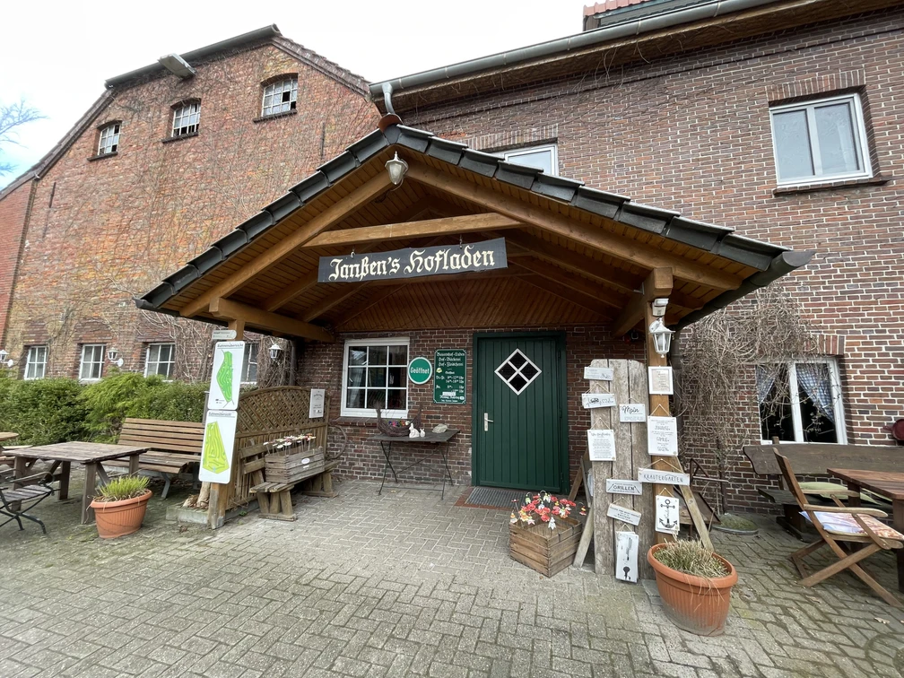 Außenansicht Hofladen Einladender Eingang zu 'Janßen's Hofladen' in einem Backsteingebäude, umgeben von Pflanzen und Schildern.