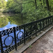 Rundteichbrücke Ein ruhiger, runder Teich von Bäumen umgeben, mit schmiedeeisernem Geländer im Vordergrund.