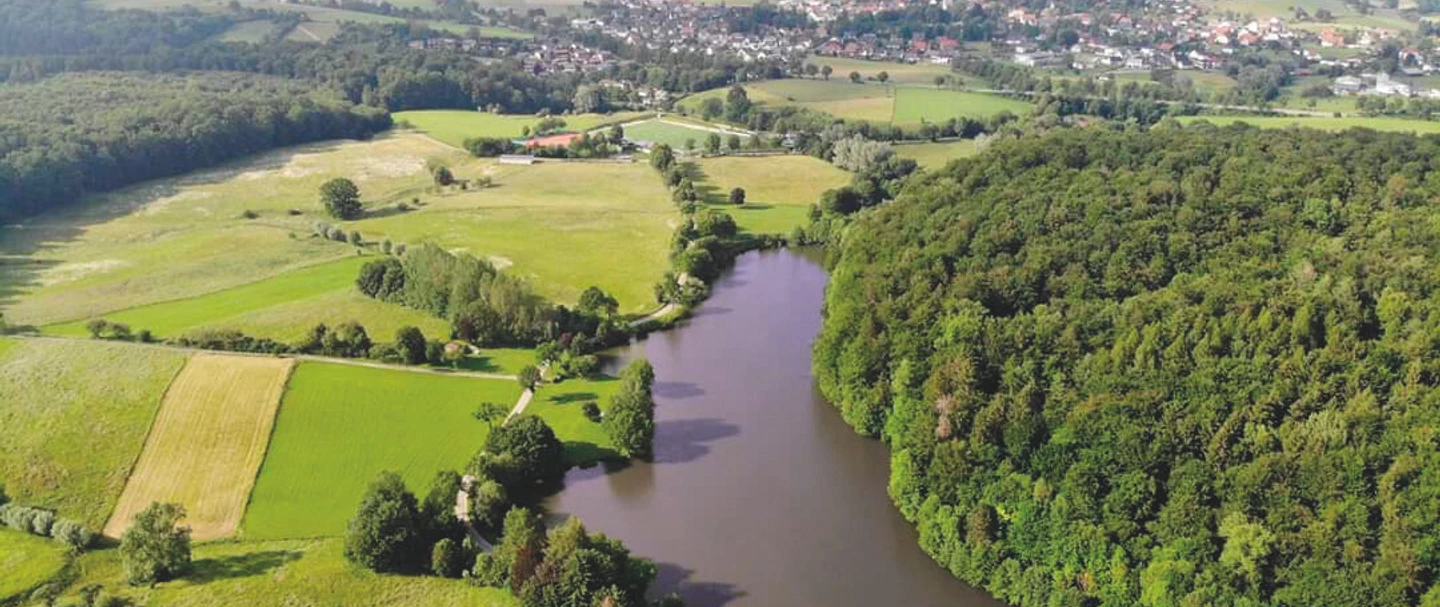 Nethestausee-Neuenheerse-2_SF_Bad-Driburger-Touristik-GmbH.png Luftaufnahme des Nethestausees, umgeben von dichten Wäldern und Feldern, Dorf im Hintergrund.
