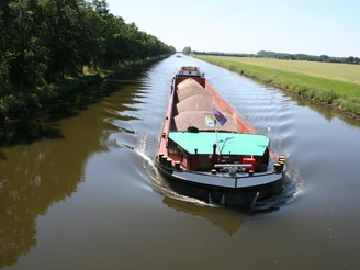 Schleusenkanal Balge Ein Frachtschiff gleitet auf einem ruhigen Kanal, umgeben von grünen Bäumen und offenen Feldern.
