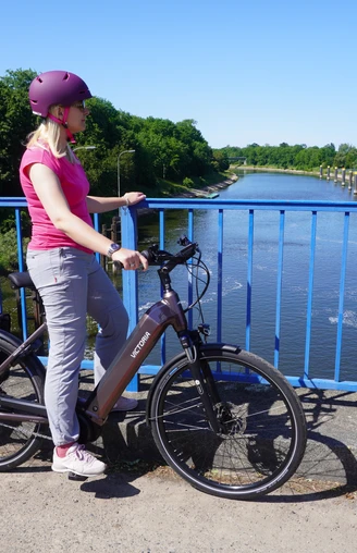 Schleusenkanal Balge Eine Radfahrerin steht auf einer blauen Brücke und blickt auf den Schleusenkanal Balge.