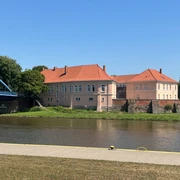 Grafenschloss Hoya Schlossanlage mit rotem Dach am Weserufer, umgeben von Bäumen, bei klarem blauem Himmel.