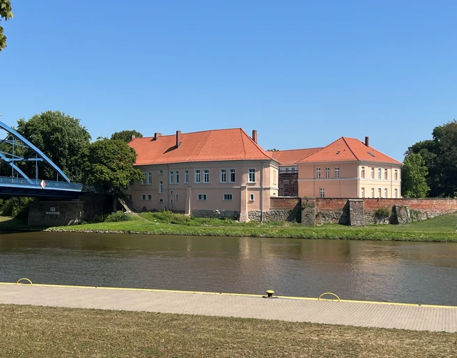 Grafenschloss Hoya Schlossanlage mit rotem Dach am Weserufer, umgeben von Bäumen, bei klarem blauem Himmel.
