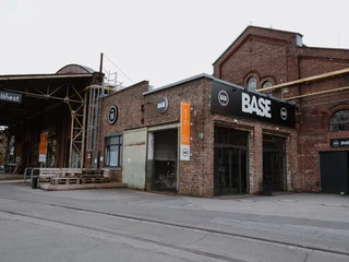 BASE draußen Das Bild zeigt die rote Backsteinfassade eines historischen Industriegebäudes mit einem großen BASE-Logo darüber. Daneben befinden sich Schilder mit der Hausnummer 1/21 und einem weiteren, kleineren Logo einer Coilfeder. Der Eingangsbereich ist modern gestaltet.The picture shows the red brick façade of a historic industrial building with a large BASE logo above it. Next to it are signs with the house number 1/21 and another, smaller logo of a coil spring. The entrance area has a modern design.