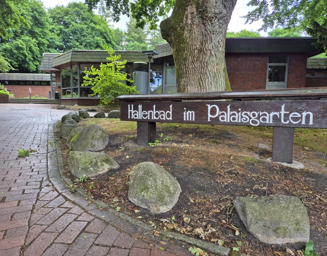 Aussenansicht Hallenbad Ein gepflegter Eingang zu einem Hallenbad im Palaisgarten, umgeben von Bäumen und einem Weg aus rötlichen Steinen.