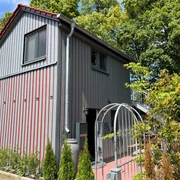 Deutsches Kreativzentrum Holz Ein zweistöckiges Gebäude aus grauen Holzbrettern, umgeben von Bäumen und einem gepflasterten Weg.