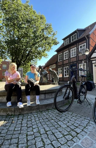 Spargelbrunnen Nienburg Zwei Frauen sitzen auf einer Mauer vor dem Spargelbrunnen in Nienburg, umgeben von Fachwerkhäusern.