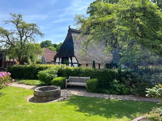 Spargelmuseum Nienburg Ein reetgedecktes Fachwerkhaus in einem grünen Garten mit einer Bank und einem alten Brunnen im Vordergrund.