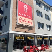 Cadera Yesterday verkleinert.jpg Außenansicht eines Cafés namens „Cadera Yesterday – Frisch von gestern“ mit großer Werbetafel an der Hauswand, beschrifteten Fenstern und roten Tischen und Stühlen auf dem Gehweg bei sonnigem Wetter.