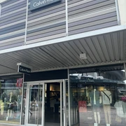 Calvin Klein.jpg Das Bild zeigt einen modernen Calvin Klein Store mit geöffneter Glastür und sichtbaren Schaufensterpuppen. Dieser lichtdurchflutete Laden befindet sich in einem eleganten Einkaufszentrum und lädt zum Stöbern ein.