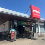 Penny Hallenbad verkleinert.jpg Eingang eines Penny-Supermarkts mit rotem Schild „PENNY. Am Hallenbad“. Vor dem Eingang stehen Einkaufswagen, Blumen auf Rollwagen und Werbeplakate. Der Himmel ist klar und blau.
