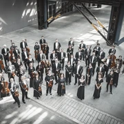 GSO_(c)Mirko Plha 3,2MB Kopie.jpg Musiker eines Orchesters stehen in einer großen Halle mit hohen Decken, die instrumente spielbereit halten.