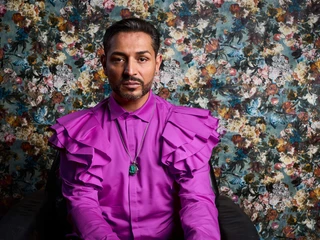 Jovanoic, Gianni Eine Person in einem leuchtend pinken Hemd mit Rüschen sitzt vor einer floral gemusterten Tapete.A person in a bright pink shirt with ruffles sits in front of a floral patterned wallpaper.