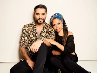 Jovanovic, Gianni Oyindamola Alashe Ein lächelnder Mann und eine Frau sitzen entspannt aneinander gelehnt vor einem neutralen Hintergrund.A smiling man and a woman sit leaning against each other in front of a neutral background.