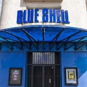 Blue Shell Das Bild zeigt den Eingangsbereich des Veranstaltungsortes „Blue Shell“ in Köln. Über dem Eingang prangt ein auffälliges, blaues Schild mit dem Schriftzug „Blue Shell“. Das Gebäude hat eine helle Fassade, wobei der Eingangsbereich in Blau gehalten ist. Ein Vordach aus Metall bietet Schutz vor Witterungseinflüssen und unterstreicht das zeitgemäße Design des Clubs.The image shows the entrance area of the venue "Blue Shell" in Cologne. Above the entryway is a prominent blue sign featuring the inscription "Blue Shell." The building has a light-colored facade, while the entry area is accented in blue. A metal canopy provides weather protection and emphasizes the contemporary design of the club.