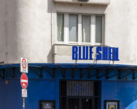 Blue Shell Die Fassade des bekannten Kölner Clubs "Blue Shell" in leuchtendem Blau mit markantem Muschel-Logo.The facade of the well-known Cologne club "Blue Shell," in vibrant blue featuring its distinctive shell logo.