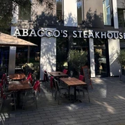 Vorderfront und Eingang des Abacco's Steakhouse mit großen Fenstern und einer Terrasse mit Stühlen und roten Decken.