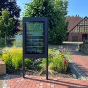 Infoterminal Winzlar Infotafel im Freien, von bunten Blumen umgeben, mit historischem Fachwerkhaus im Hintergrund.