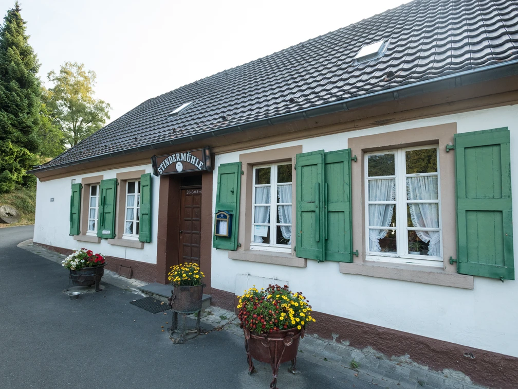 Stindermühle in Erkrath.jpg Weißes Gebäude mit grünen Fensterläden und einem Schild "Stindermühle", flankiert von Blumenkübeln.