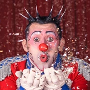 Great Christmas Circus Frankfurt - Henry the prince of clowns Clown in bunter Uniform pustet glitzernden Konfetti vor rotem Vorhang.Clown in a colorful uniform blows glittering confetti in front of a red curtain.