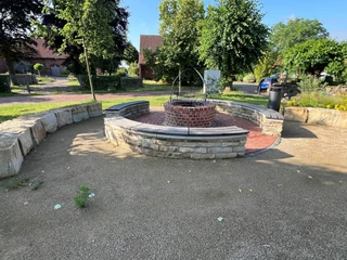 Eierbratstelle Winzlar Halbrunder, mit Steinen eingefasster Sitzbereich mit einem zentralen, steinernen Brunnen in sonniger Umgebung.