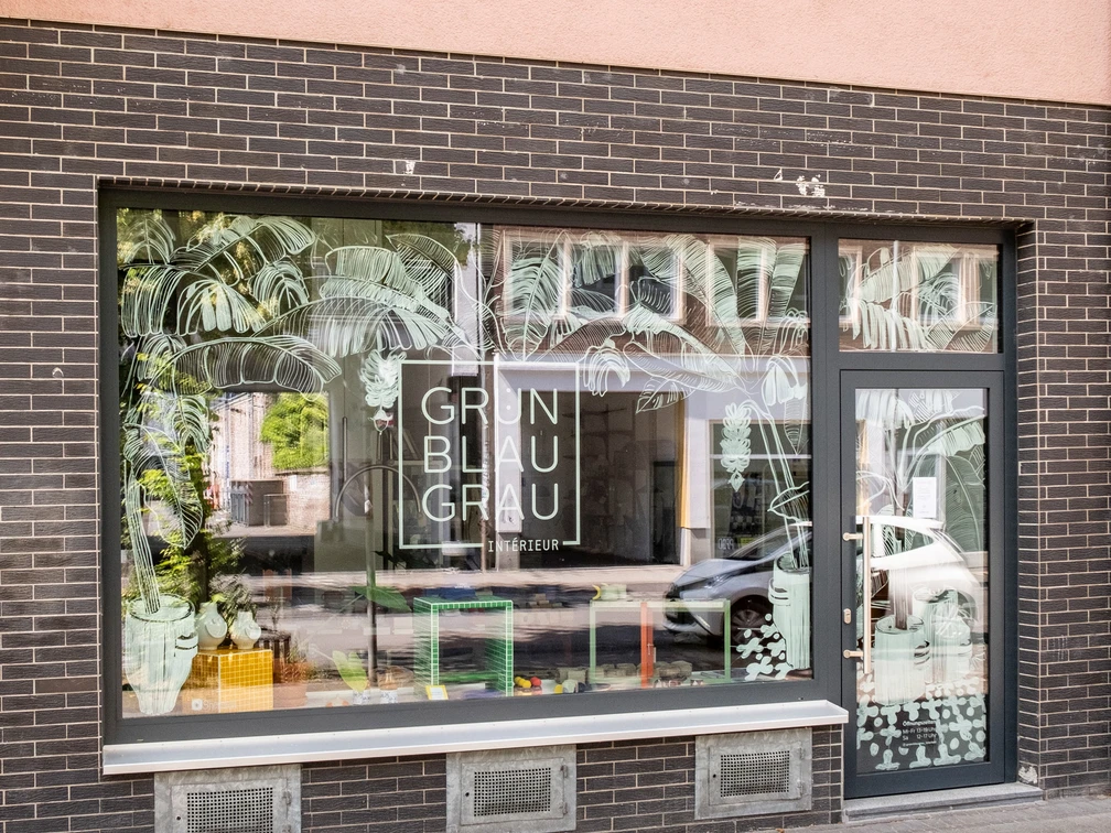 GrünBlauGrau Intérieur Die Fassade eines Geschäfts mit großen Fenstern zeigt den Schriftzug "GrünBlauGrau Intérieur" vor einem dekorierten Schaufenster.The façade of a store with large windows shows the lettering "GrünBlauGrau Intérieur" in front of a decorated shop window.