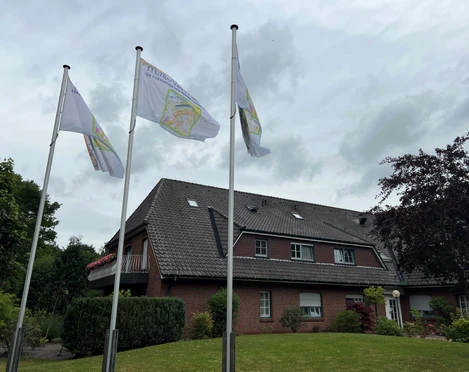Außenansicht.jpg Gemütliches Backsteinhäuser mit drei wehenden Flaggen, umgeben von gepflegtem Grün unter grauem Himmel.